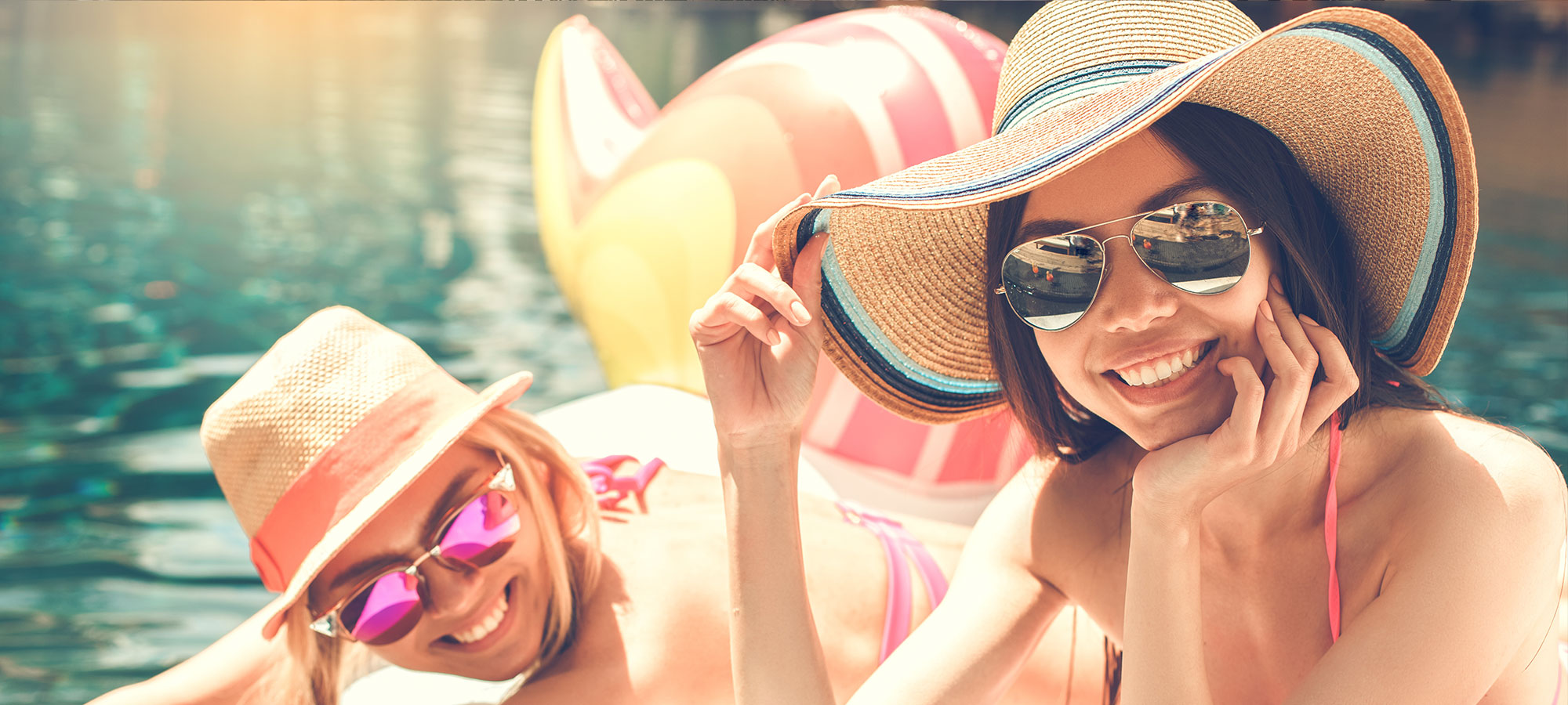 Two women wearing shades and straw hat smiling by the poolside