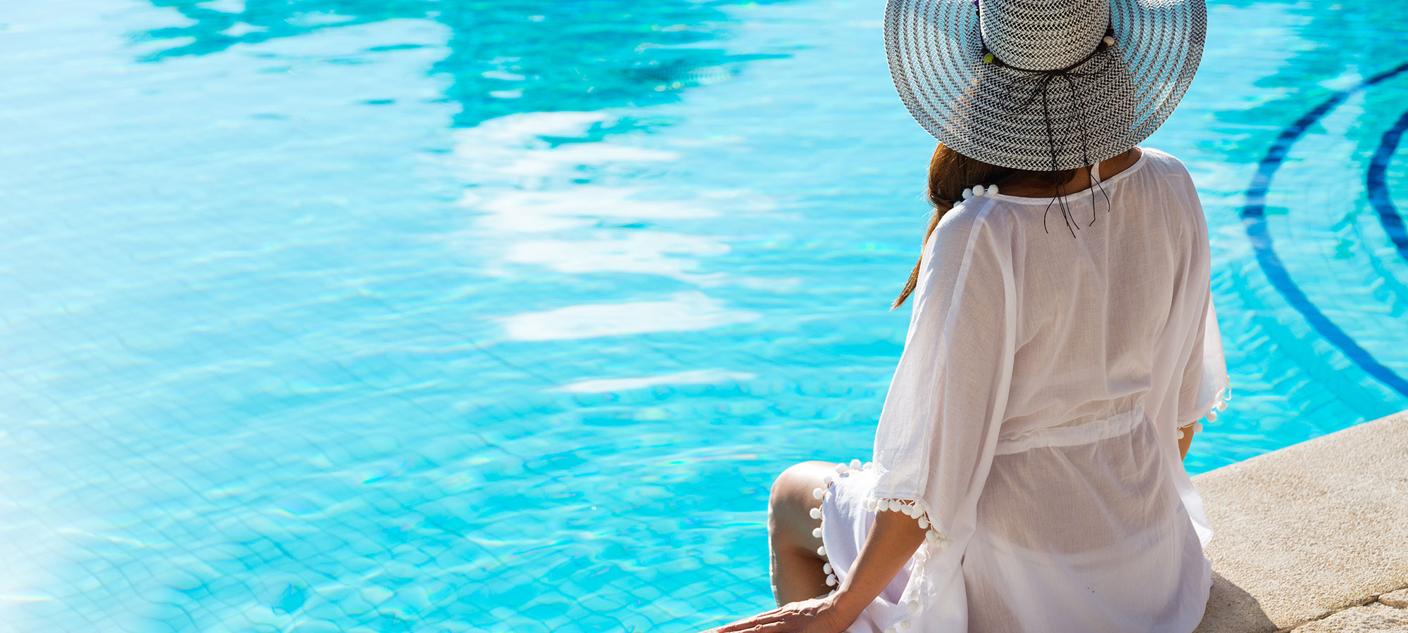 Woman wearing a white gown and straw hat sitting by pool