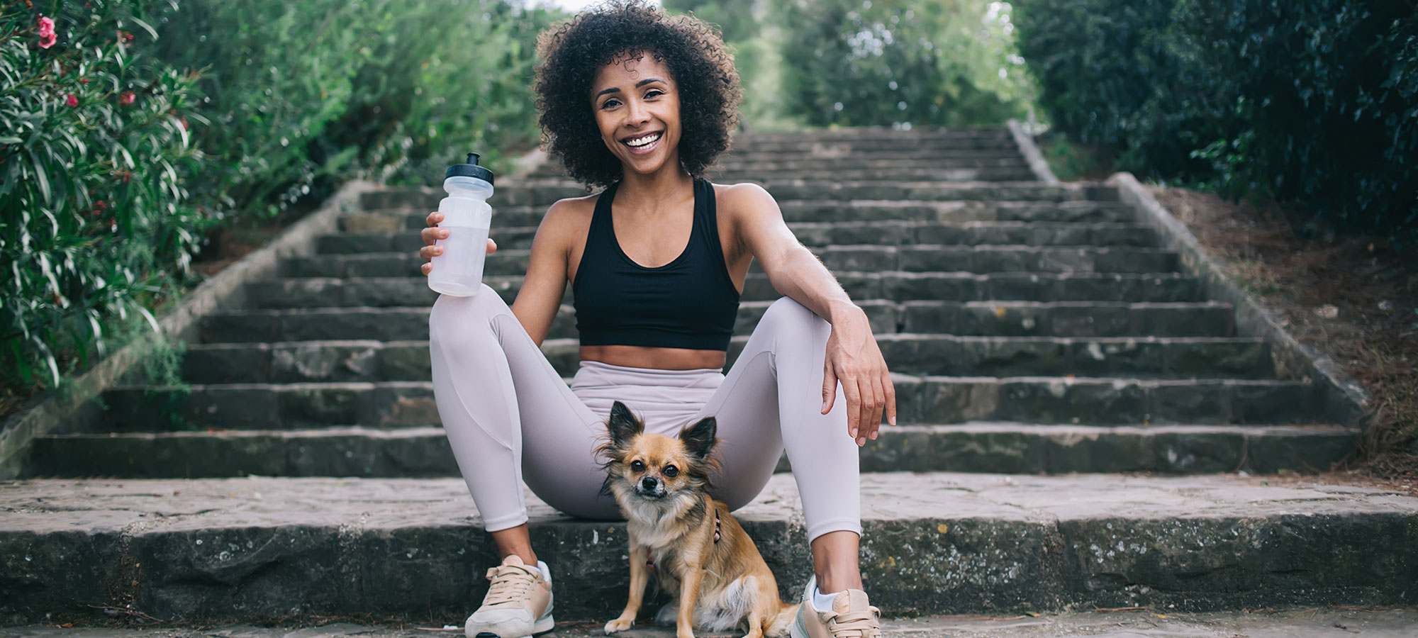 Woman sitting on concrete stairs with dog holding water bottle and smiling