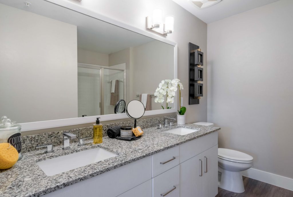 Interior bathroom with dual sink, large mirror, designer lighting, wood-style flooring, and granite countertops